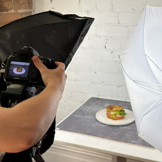 A person holding a DSLR camera with a softbox light on a tripod, pointing it toward a small plate of food on a table in a studio setup with a white umbrella light.